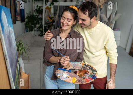 Loving couple of artists enjoys finished painting, partners pleased with result and share happiness. Stock Photo