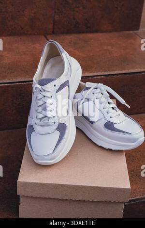 Minimalist eco-friendly white sneakers placed on recycled cardboard box, clean design for sustainable fashion concepts. New unbranded white sneakers footwear mockups and modern product displays. For branding, e-commerce and packaging design Stock Photo