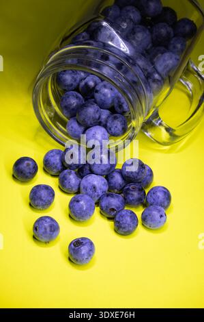 Fresh blueberries spilling from an overturned clear glass jar onto a vibrant yellow background creating a bold color contrast between deep blue Stock Photo