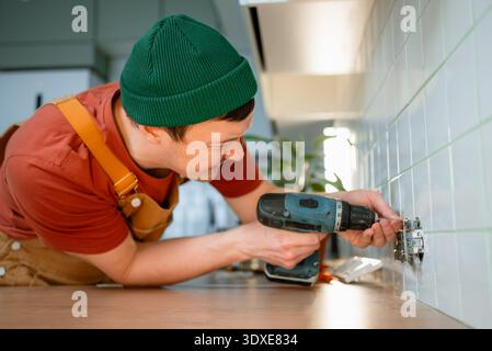 Handyman wearing a green beanie and overalls installing an electrical outlet into a tiled kitchen wall countertop, focusing on the task with a cordless drill Stock Photo