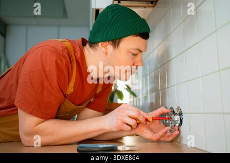 Man repairs wall socket with screwdriver, performing electrical work and home renovation tasks in a modern kitchen, focused handyman providing professional maintenance services Stock Photo