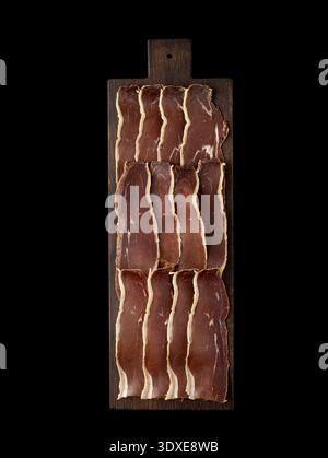 Thinly sliced meat, including jamon, artistically arranged on a rustic dark wooden cutting board. Isolated on a black background, top view. Stock Photo