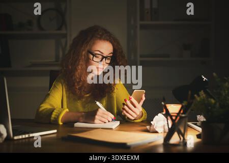 Woman Working Late at Night in Home Office While Using Phone and Taking Notes Stock Photo