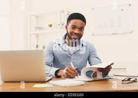 Man Works at Desk With Laptop While Writing Notes and Smiling in Modern Office Setting Stock Photo