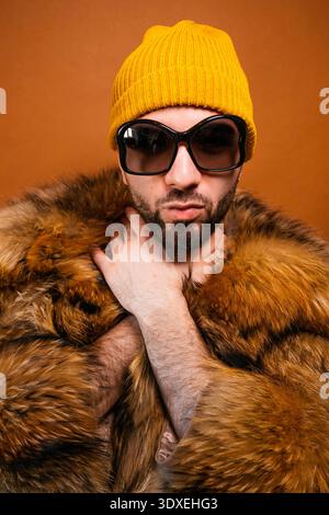 Young adult man wearing a fur coat, yellow beanie, and sunglasses against a brown background Stock Photo