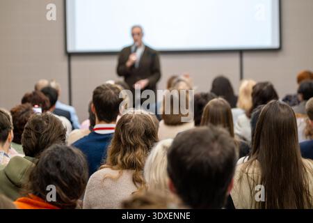 Crowd of professionals listening to presenter during business conference, some attendees recording slides with smartphones. Stock Photo
