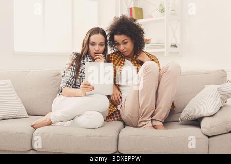 Two Friends Sit Together on a Couch While Looking at a Tablet and Sharing an Experience Stock Photo
