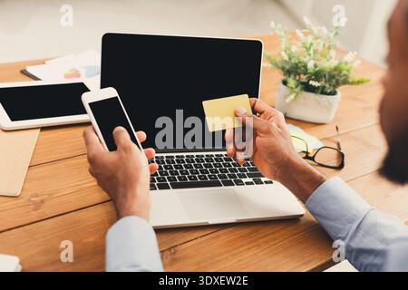 Man Uses Mobile Phone and Card While Sitting at a Desk With a Laptop and Tablet Stock Photo