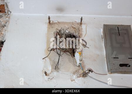 A close-up of a damaged wall section featuring a chaotic nest of old electrical wires, a single circuit breaker, and an empty metal fuse box during a Stock Photo