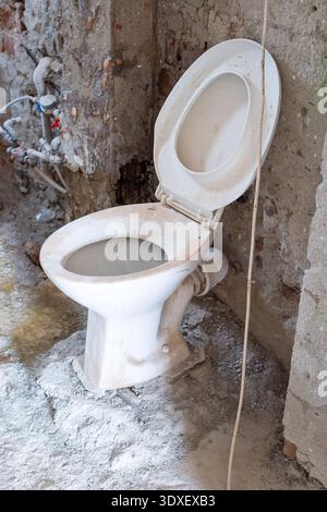 A dusty white ceramic toilet sitting in a construction site, surrounded by raw concrete floors, exposed brick walls, and unfinished plumbing pipes Stock Photo