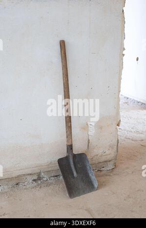 Description: A weathered metal spade with a wooden handle rests against an unfinished plaster wall on a dusty concrete construction site floor during Stock Photo