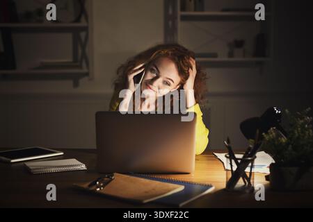 Woman Sitting at Desk Talking on Phone While Using Laptop in Dark Room Stock Photo