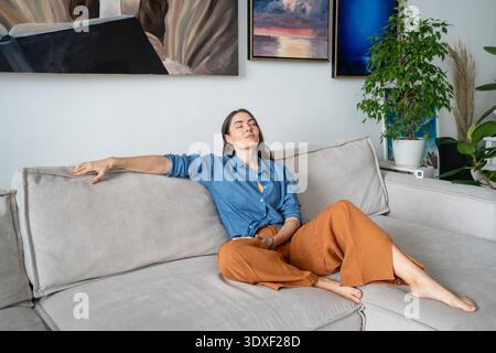 Dreamy tranquil woman artist sits on couch at home gallery apartment, thinking about artwork ideas. Stock Photo