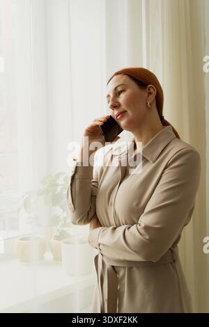 Woman is having a business conversation, standing by the window with her smartphone, feeling thoughtful and socializing while working from home Stock Photo