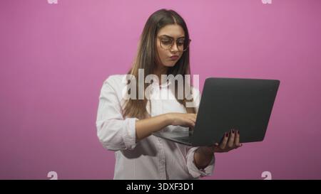 Woman holding laptop and typing with hands in a pink studio, looking at screen with pursed lips; concentration remote work productivity. Stock Photo