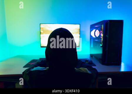 Silhouette of gamer wearing hoodie while playing racing video game on bright monitor with colorful LED background Stock Photo