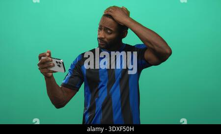 Man holding smartphone with hand on forehead in green studio set wearing blue soccer jersey and looking at screen; frustration technical issue. Stock Photo