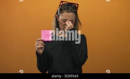 Young blonde woman pinches nose with thumb and forefinger while holding pink sticky note with smile drawing against orange studio wall, wearing black Stock Photo