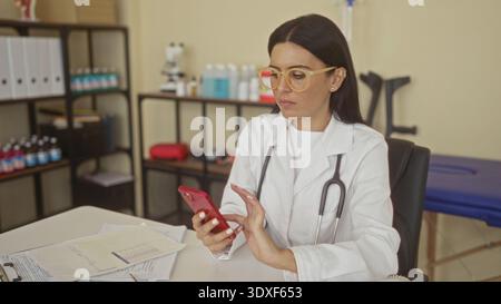 Woman doctor with stethoscope taps red smartphone with index finger at exam desk in building; focused professional care. Stock Photo