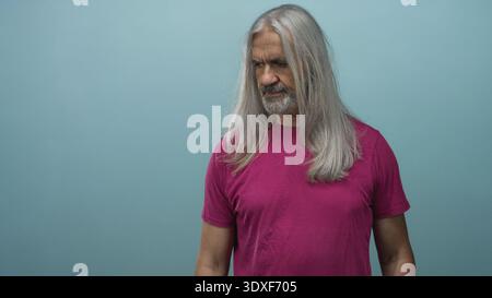 Middle aged man with long gray hair wearing magenta tee in teal studio looking sideways and talking with expressive face; surprise reflection. Stock Photo