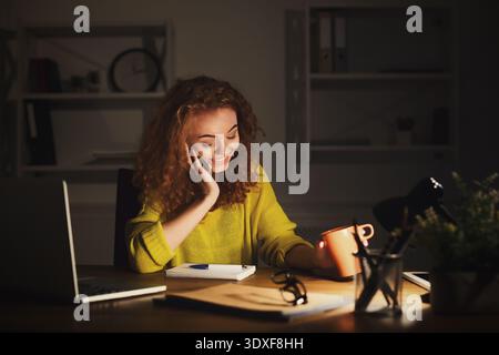 Young Woman Smiles While Working Late at Night in a Cozy Home Office Setting With a Warm Drink Stock Photo