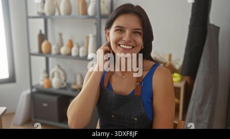 Woman adjusts hair with a smile while wearing apron in studio surrounded by pottery vases on shelves; creative joy. Stock Photo