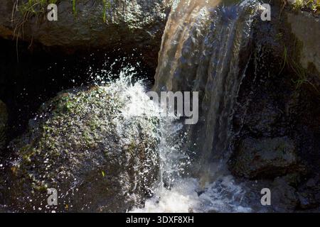 A dynamic close-up of a small waterfall. Murky water flows over dark, moss-covered rocks, creating a bright splash. High-contrast lighting highlights Stock Photo