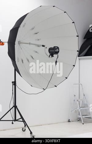 Professional photography studio setup. A large parabolic umbrella softbox with a strobe flash is on a stand in a minimalist white room. Behind the sce Stock Photo