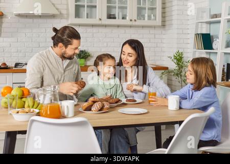 Happy family of four having breakfast together in modern kitchen, enjoying fresh pastries and juice. Stock Photo