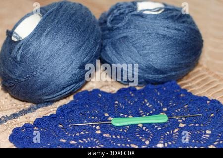 Close-up still life of crochet supplies. A green hook lies on a handmade blue lace doily, with two balls of yarn in the background. Evokes creativity, Stock Photo