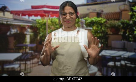 Woman in apron raises hands showing palms in cafe building patio, wearing glasses and sleeveless top, slight grimace; concern hospitality. Stock Photo