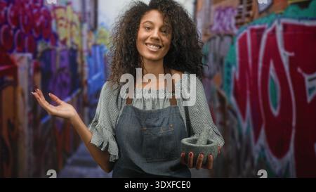 Teen woman with curly hair wearing apron holds mortar and pestle in left hand and shows right palm up gesture while smiling in graffiti street; urban Stock Photo