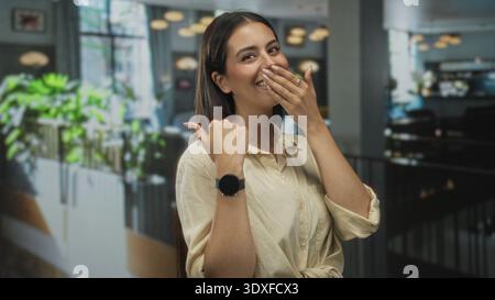 Young brunette woman covering mouth with hand and thumb pointing to herself in restaurant building; surprise playful moment. Stock Photo