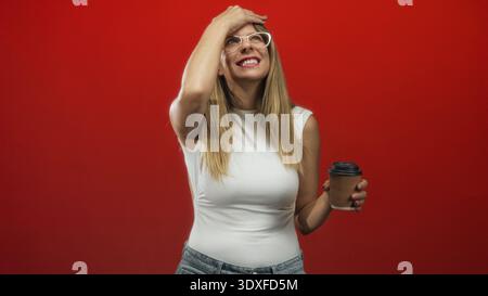 Woman in white tank top and jeans wearing glasses holding corrugated paper coffee cup with hand on forehead gesture in studio; embarrassed smile. Stock Photo