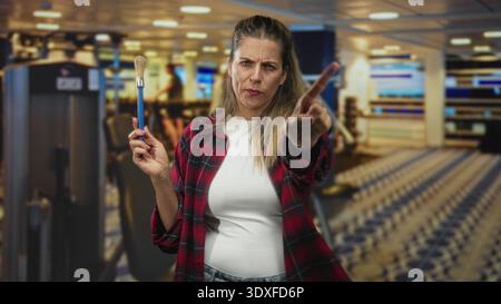 Woman painter holding paintbrush and touching chin while studying bristles in building filled with gym machines and patterned floor; creative contempl Stock Photo