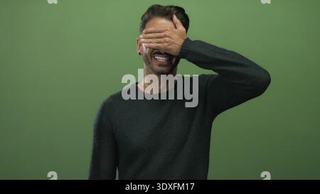 Young bearded man covers eyes with hand while smiling in studio; shyness mystery privacy playfulness. Stock Photo