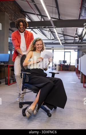 Diverse coworkers pushing rolling chair down aisle in open plan office holding printed papers. Collaboration, teamwork, modern, vibrant, casual, productivity, dynamic || Model released Stock Photo