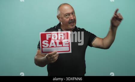 Senior hispanic man holds for sale sign with right hand and looks to side wearing black polo in studio; home sale hope. Stock Photo