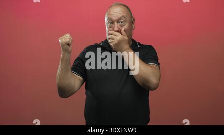 Hispanic senior man covers mouth with hand, clenches fist and looks startled in red studio; surprise silence. Stock Photo