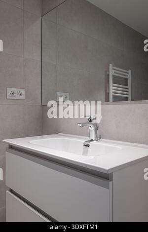 Modern bathroom interior with a sleek white sink, chrome faucet, and gray tiled walls. A mirror reflects the minimalist design and a towel radiator is Stock Photo