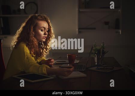 Student Works on Project at Home Late in the Evening While Using a Laptop and Taking Notes Stock Photo