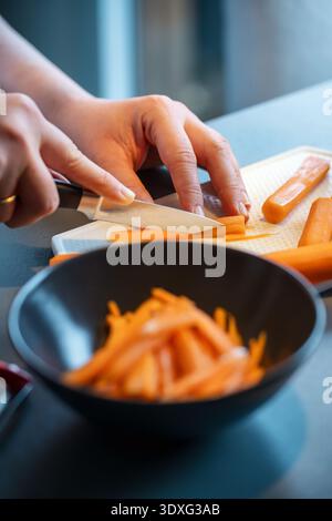 Close-up of hands cutting a carrot into thin strips on a white cutting board. A dark bowl with sliced carrot pieces is visible in the foreground. Stock Photo