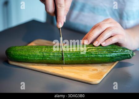 Close-up of hands cutting a fresh green cucumber in half on a wooden cutting board. Stock Photo