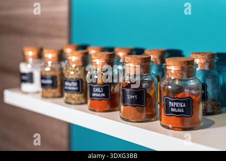 Glass spice jars with cork lids arranged on a white kitchen shelf. The jar labeled cinnamon in German is in focus. Stock Photo