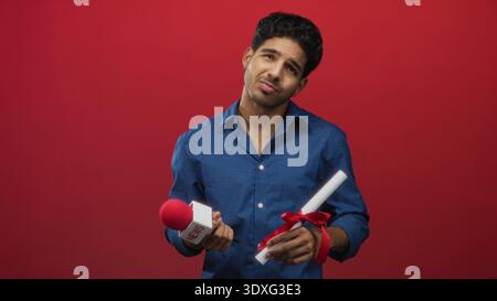 Man holding red news microphone and ribboned diploma with hands visible against a red backdrop in studio; thoughtful contemplation. Stock Photo