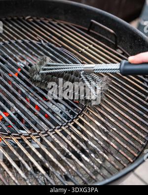 Close-up detail of a person cleaning a charcoal grill grate with a wire brush over glowing coals. Stock Photo