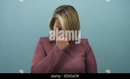 Blonde woman pinches nose wearing a mauve top in a light blue studio set reacting to a foul smell; disgust. Stock Photo