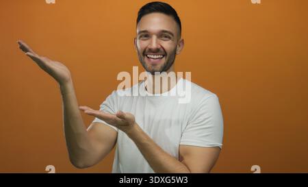 Young hispanic man smiling and extending arms to present something against a bright orange background, showcasing a welcoming gesture with joyful expr Stock Photo