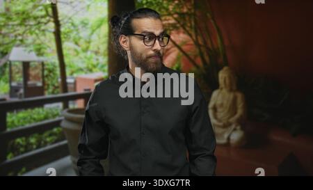 Man with glasses and beard standing in a hotel building lobby, wearing black shirt and gazing sideways near balcony and statue; contemplation. Stock Photo