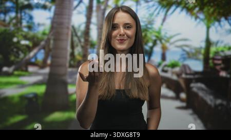 Young hispanic woman with long brunette hair extends palm, beckons with open hand wearing black strapless top in studio; welcoming warmth. Stock Photo
